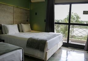 Quarto de hotel com cama, mesa de cabeceira, janela com vista para o verde e varanda com grade.