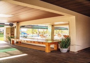 Uma área de estar coberta ao ar livre, com um banco de madeira, plantas em vasos e vista para uma piscina.