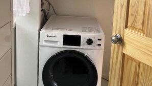 A compact, white front-loading washing machine in a small laundry room with a wooden door and a shelf above it holding folded towels. - Mini Mod Apartment, Private- Parking (Winooski)