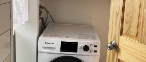 Une petite machine à laver blanche à chargement frontal, installée dans un petit espace buanderie avec une porte en bois et une étagère au-dessus, sur laquelle sont pliés des serviettes.