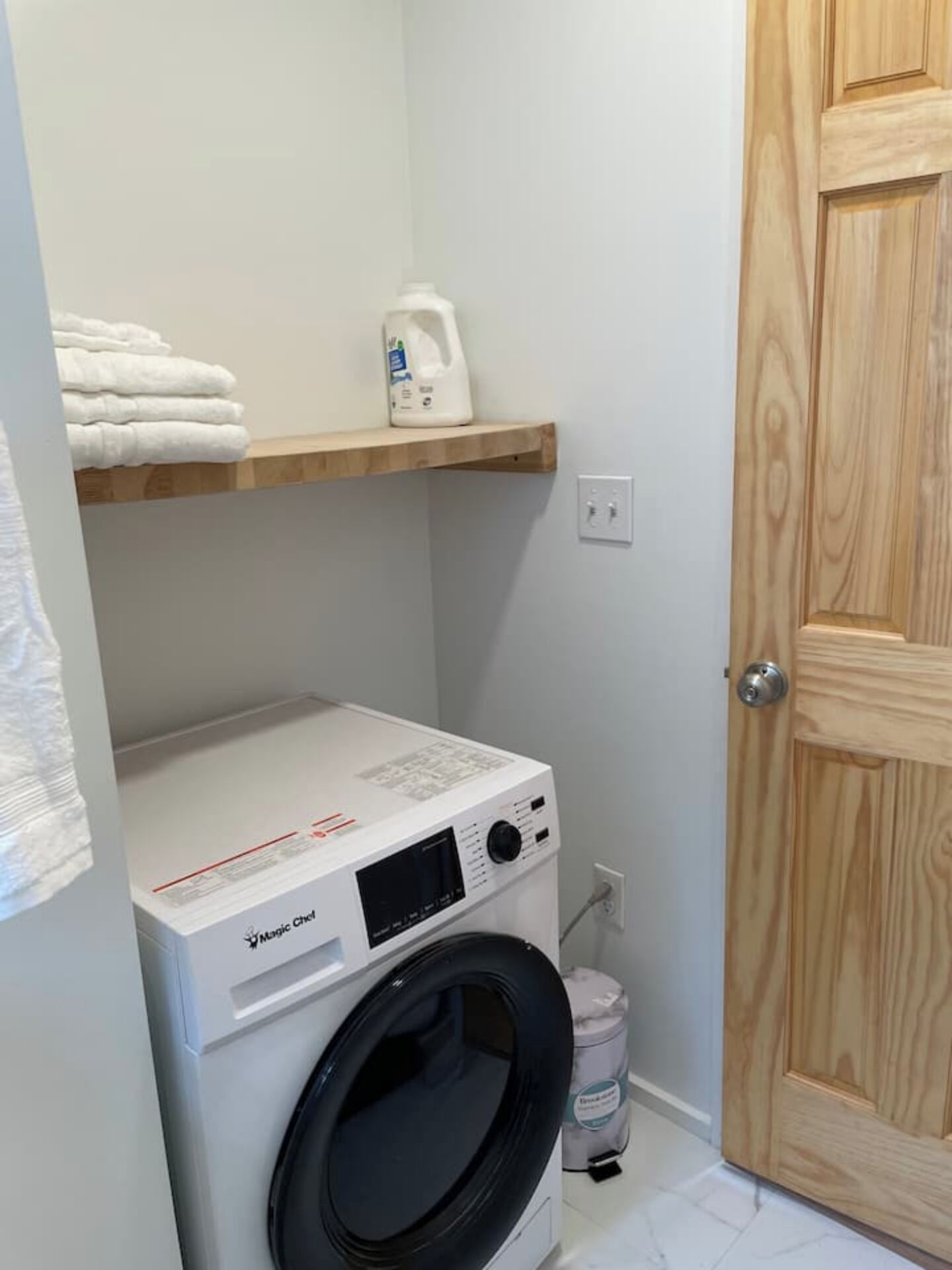 A compact laundry area with a front-loading washing machine, a shelf with folded towels, and a door.