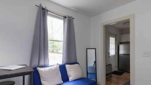 A room with a blue sofa, a small table with a chair, a mirror, and a kitchen area visible through an open door. - Mini Mod Apartment, Private- Parking (Winooski)