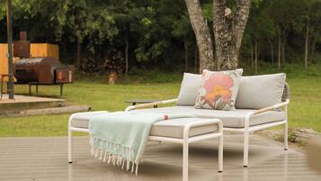 A white metal sofa with a light green throw blanket and two pillows with floral designs, positioned on a wooden deck with a grassy area and trees in the background.