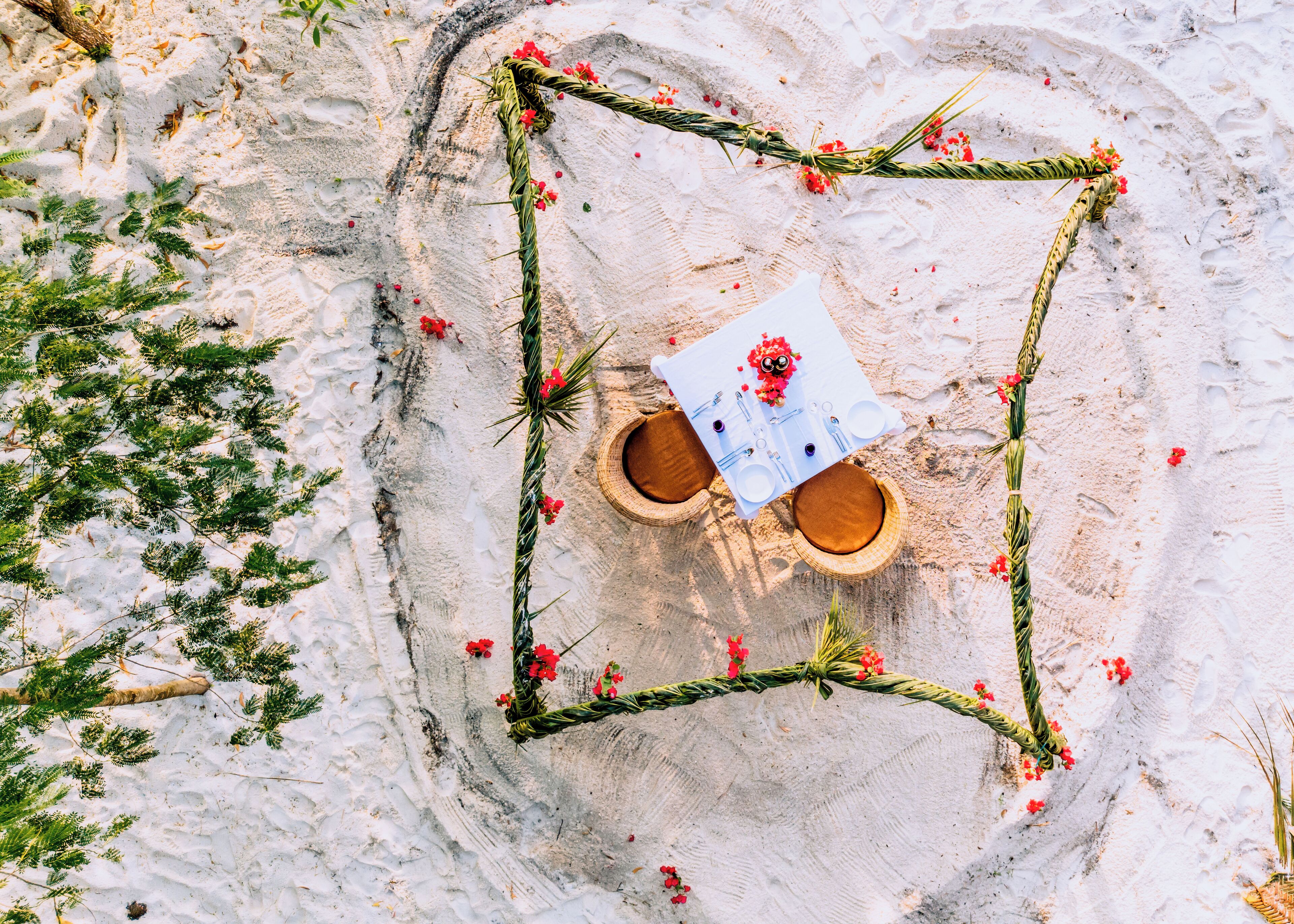 An aerial view of a square table set up on a sandy surface with a white tablecloth, surrounded by greenery and red flowers.