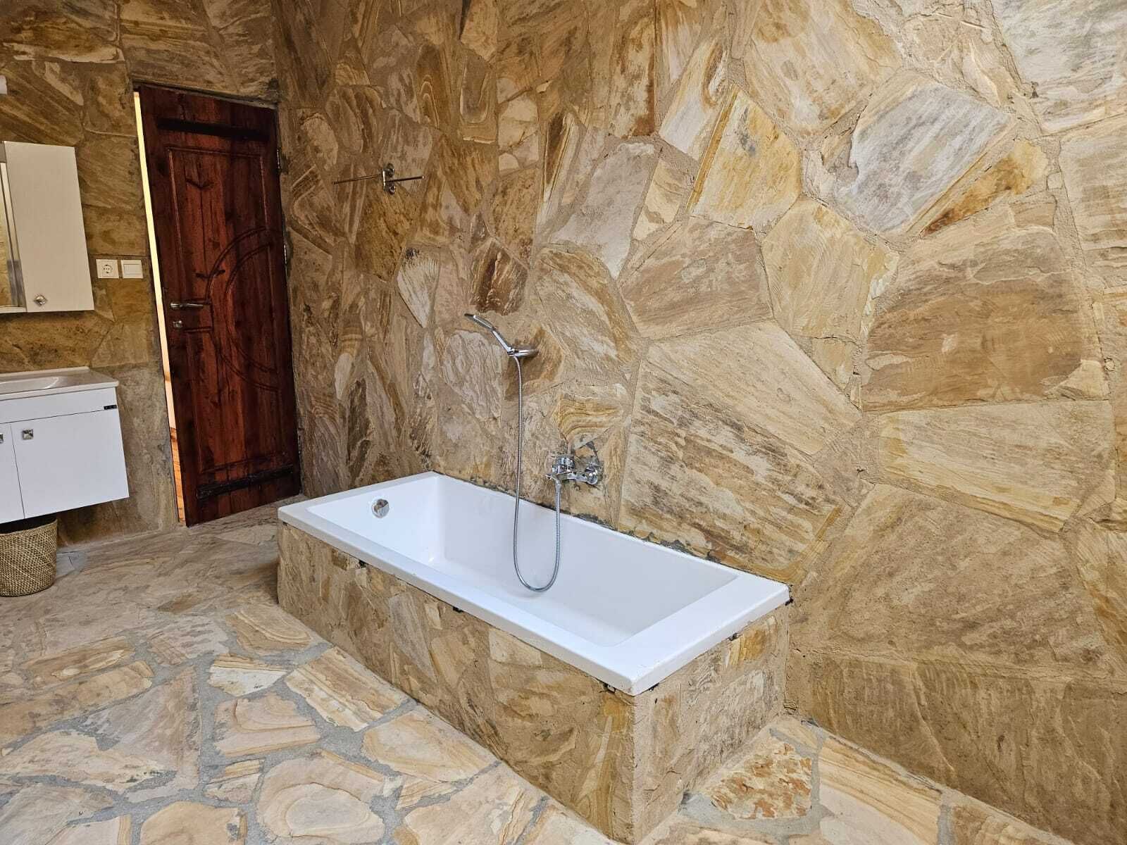 Casa de banho com parede de pedra, porta de madeira, banheira branca e lavatório branco com armário.