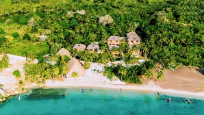 Vista aérea de un complejo turístico costero con varias construcciones tipo bungalow rodeadas de exuberante vegetación y una playa de arena.