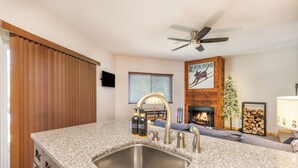 A modern kitchen with granite countertops, stainless steel sink, and a built-in stove. There is a wooden ceiling fan, a fireplace, and a framed picture on the wall.