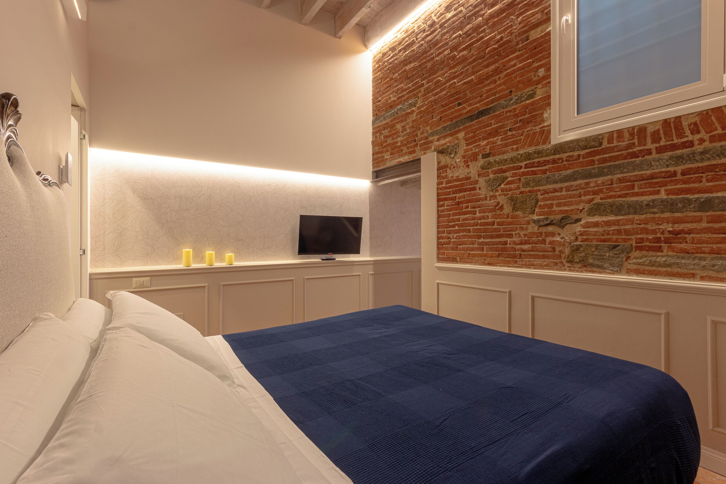 A bedroom with a bed, a flat-screen TV, and a brick wall.
