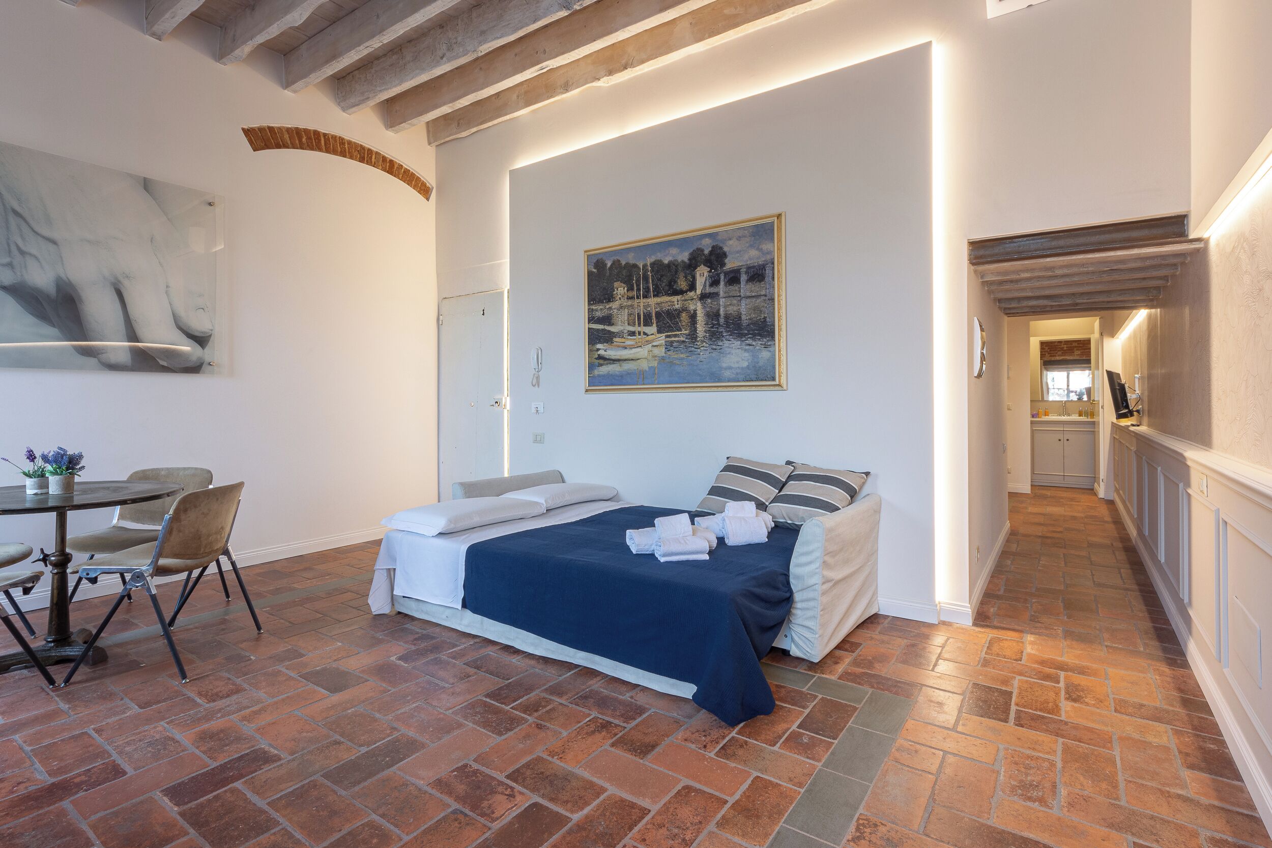 A bedroom with a bed, a painting, a dining table with chairs, and a hallway.