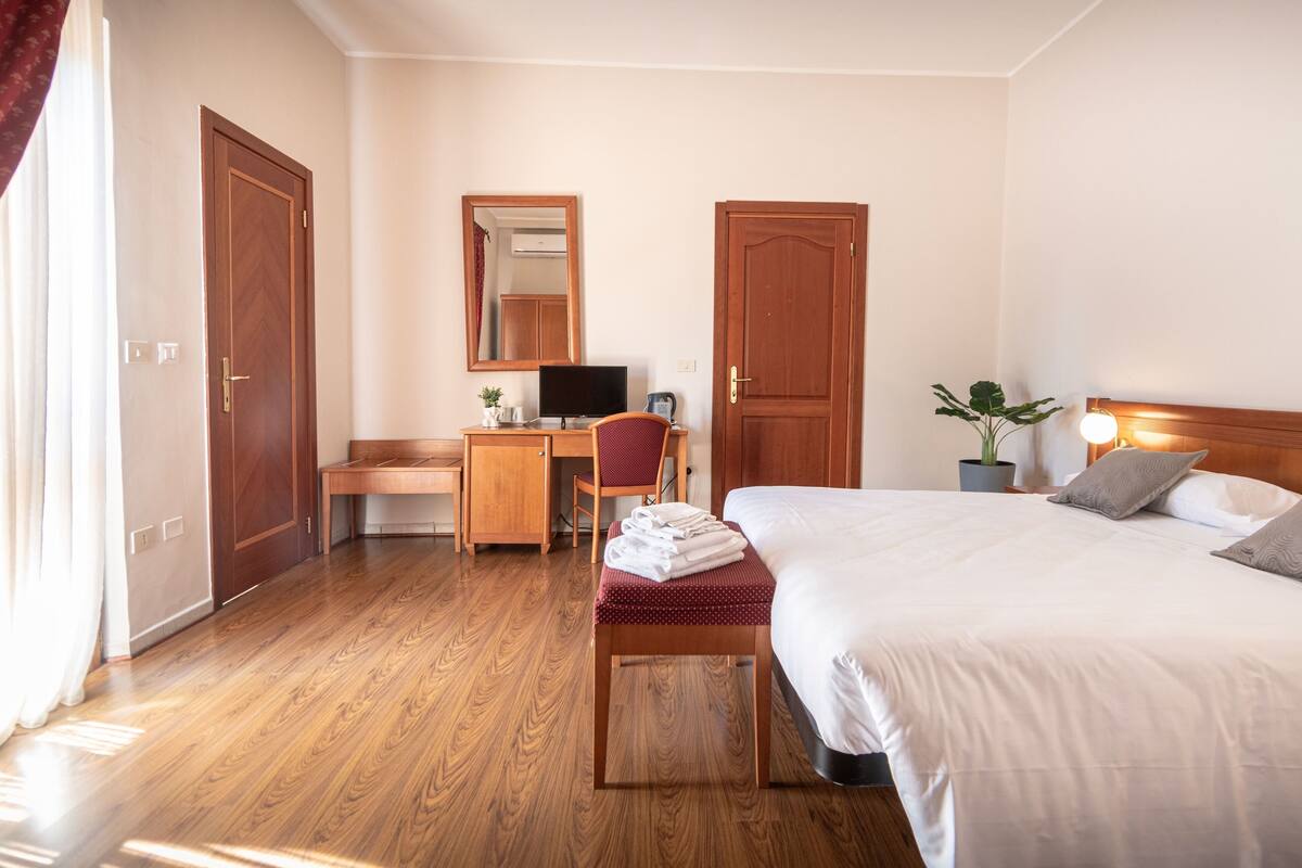 A hotel room with a wooden floor, a desk with a chair, a bed with pillows, and a potted plant.