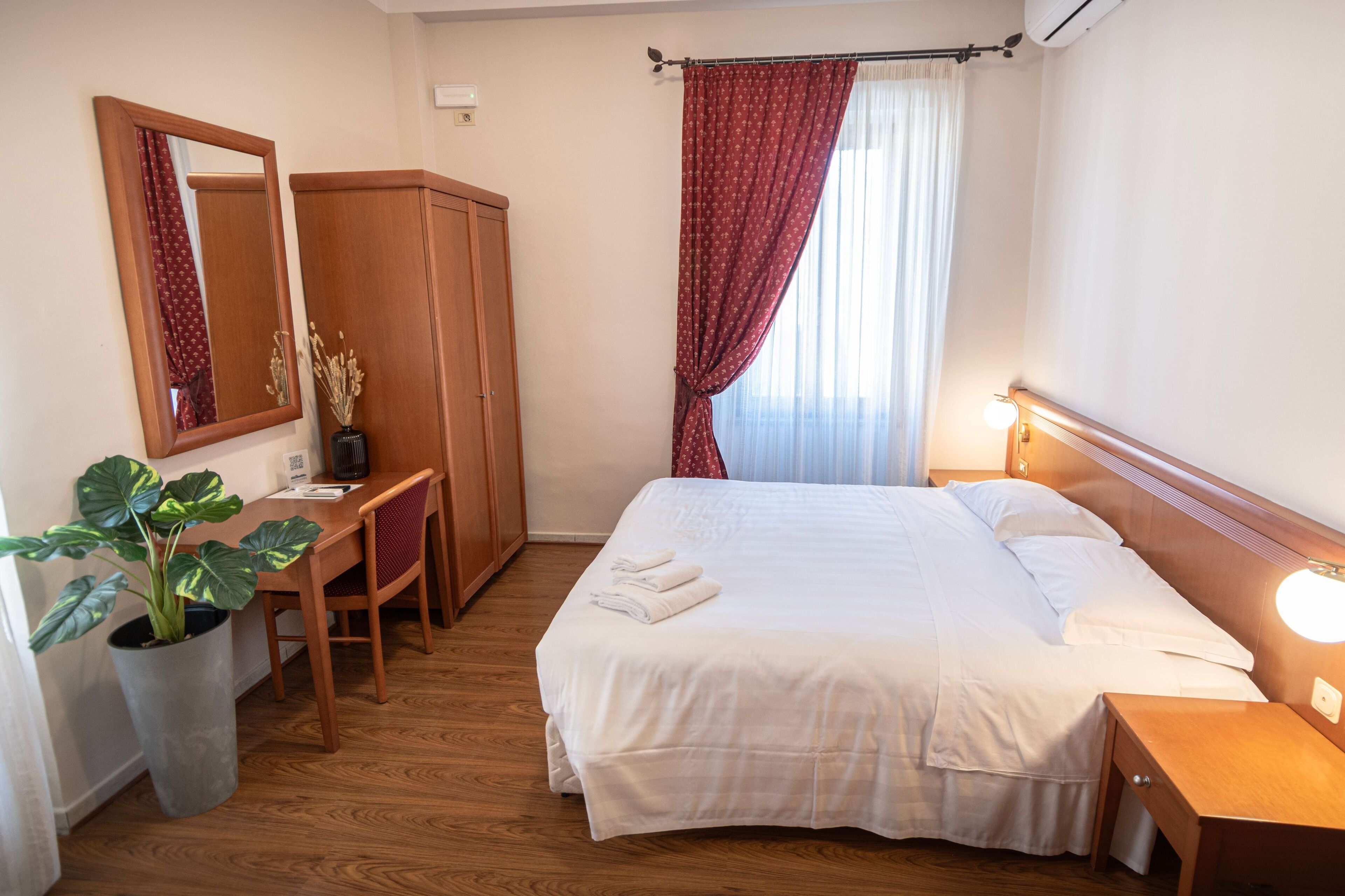 A hotel room with a bed, a desk, a chair, a wardrobe, and a potted plant.