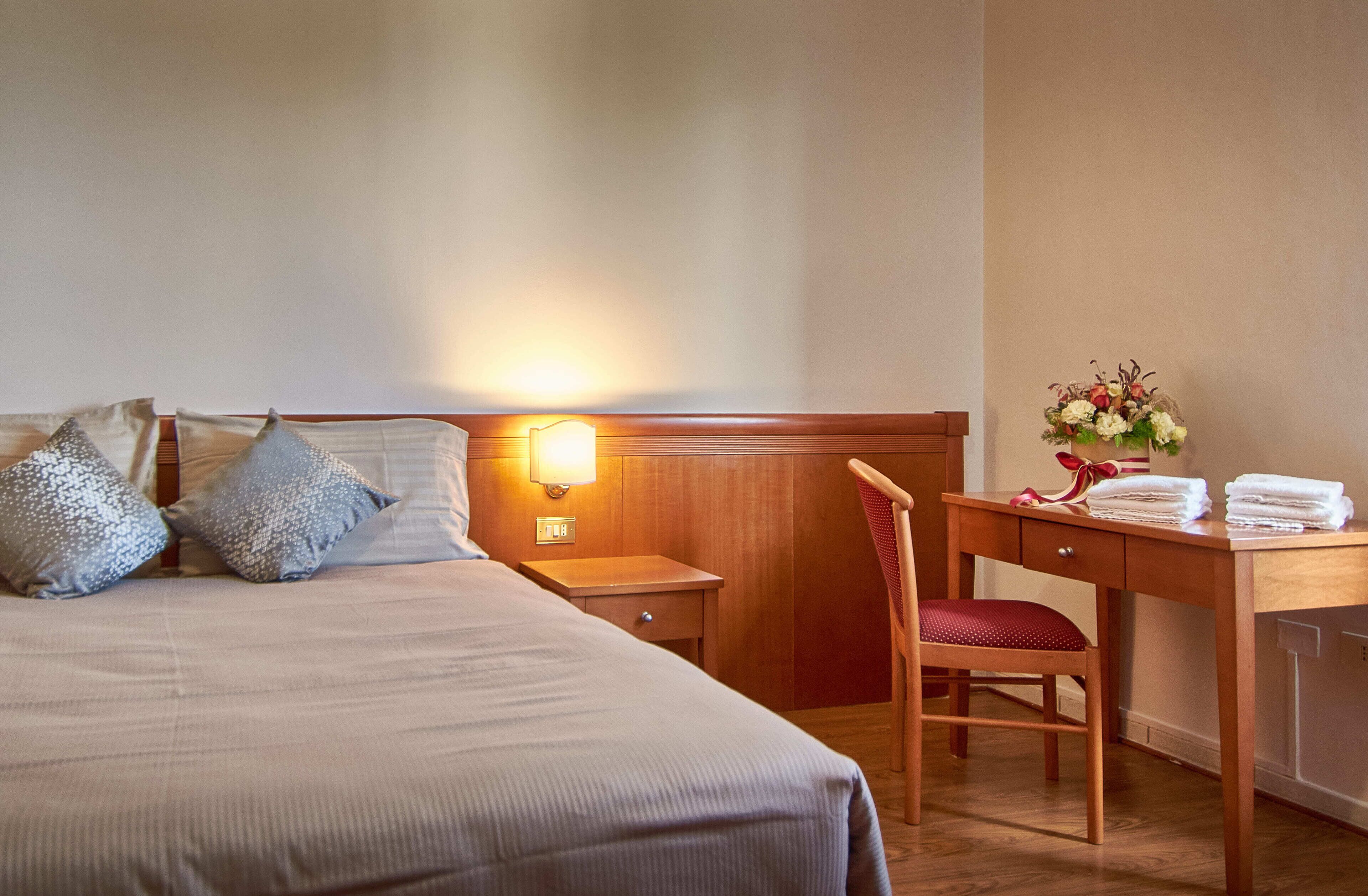Quarto de hotel com cama, escrivaninha, cadeira, mesa de cabeceira e luminária na parede.