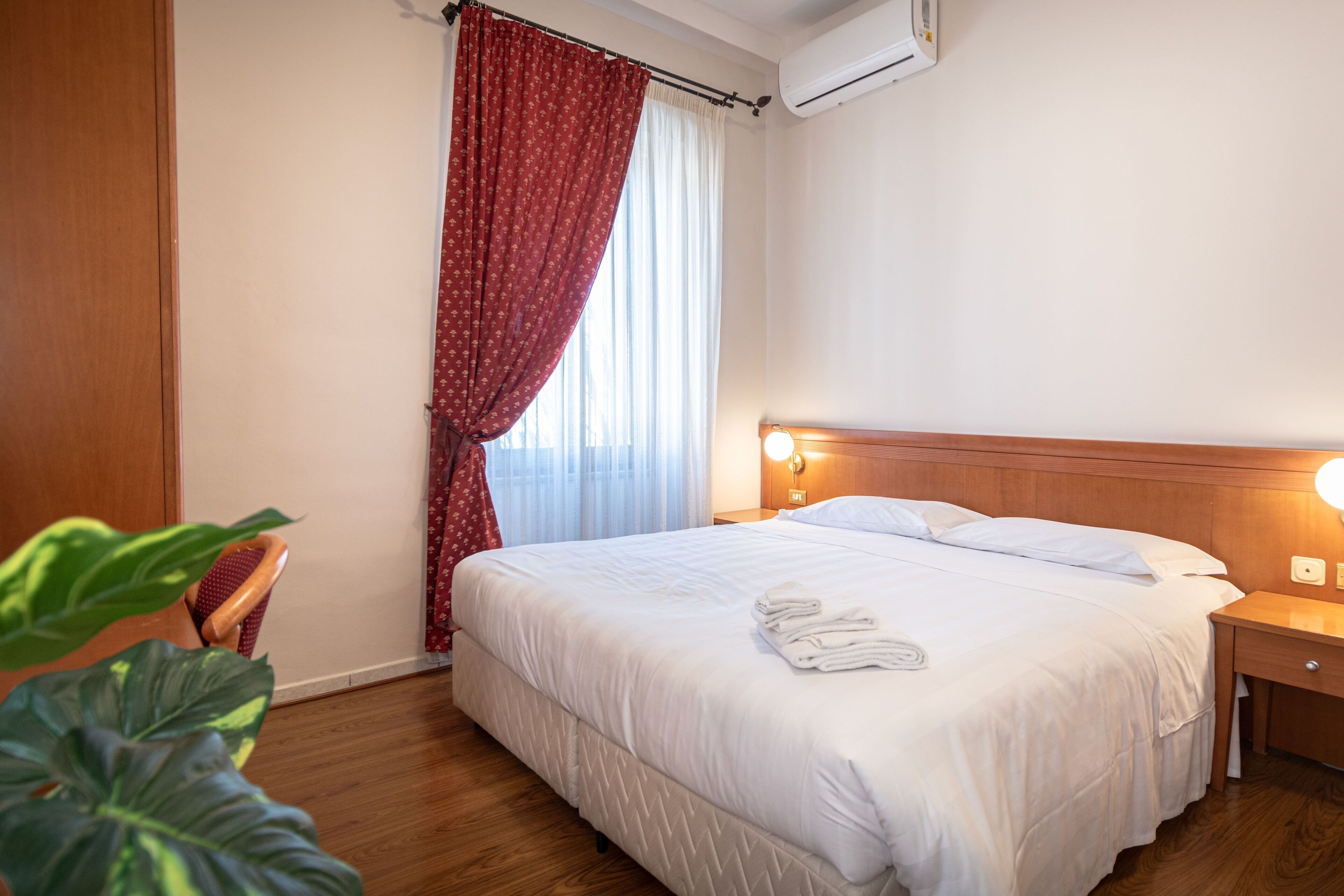 Een hotelkamer met een bed, nachtkastje, houten hoofdbord, raam met gordijnen en een bloempot met plant.