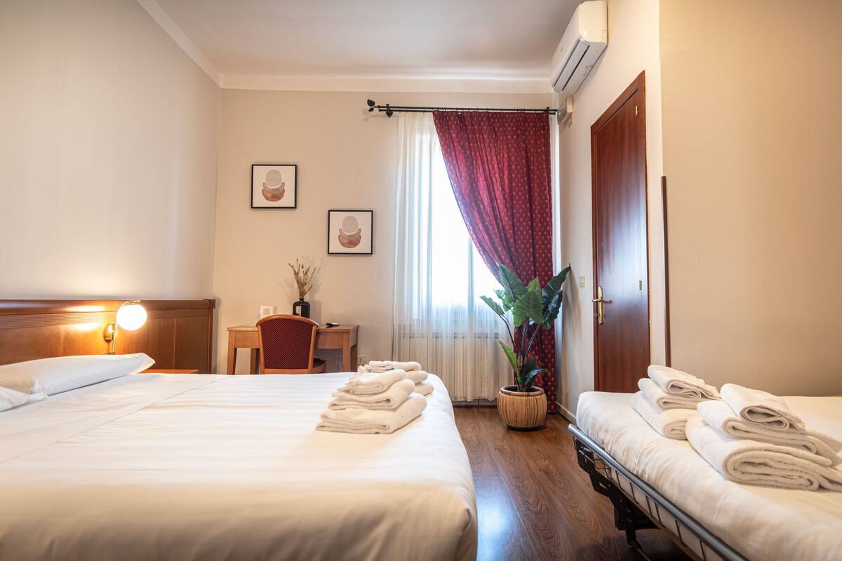 A hotel room with two beds, a desk, a chair, a potted plant, and a window with curtains.