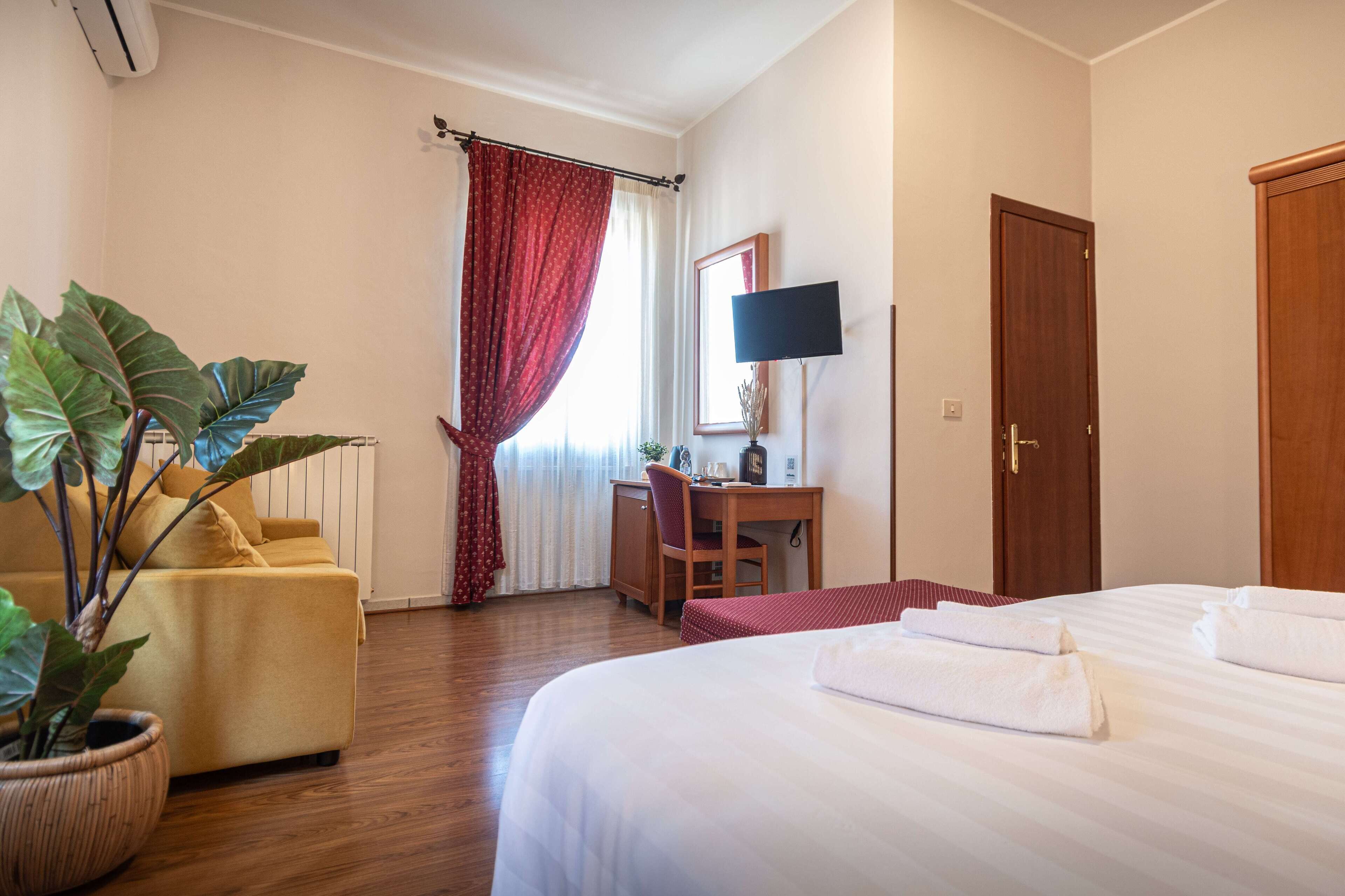 A hotel room with a bed, a desk, a chair, a potted plant, a TV, and a door.