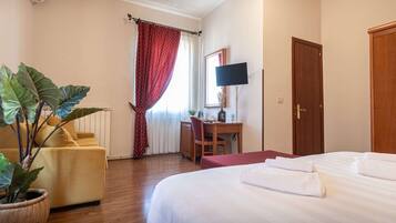 A hotel room with a bed, a desk, a chair, a potted plant, a TV, and a door.
