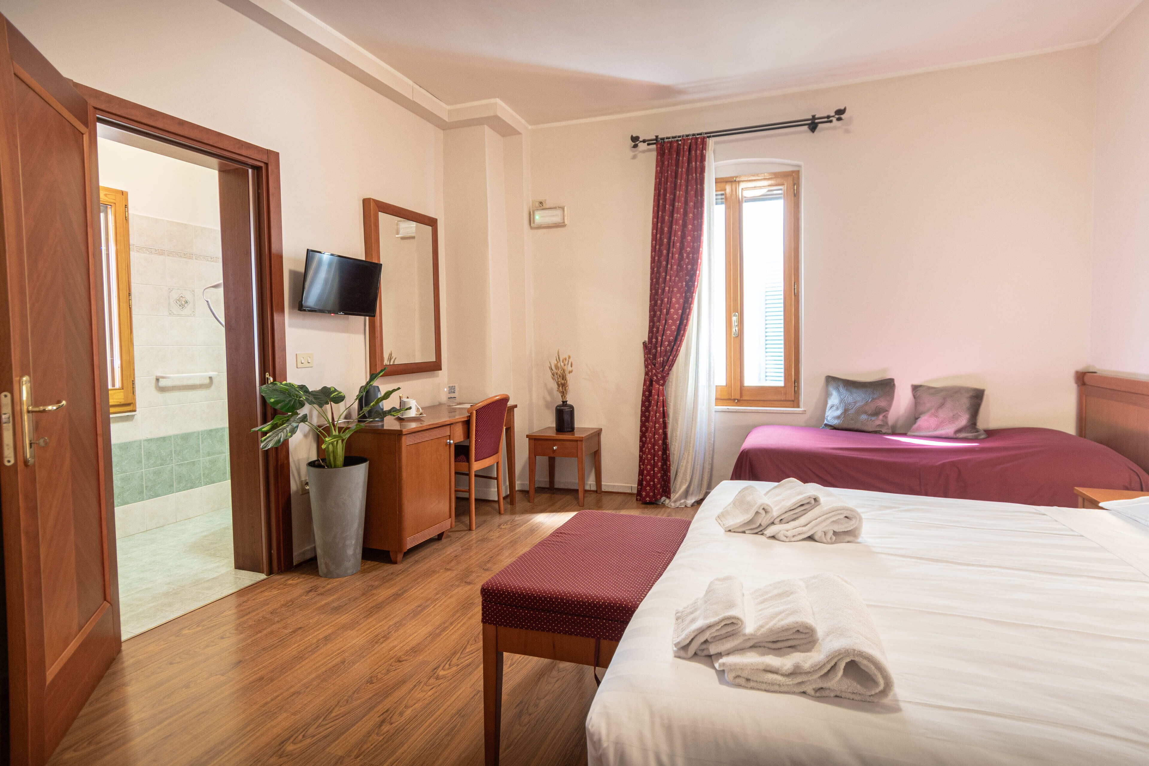 Une chambre d’hôtel avec un plancher en bois, un bureau avec une chaise, un téléviseur fixé au mur, un lit avec deux oreillers et une fenêtre avec des rideaux.