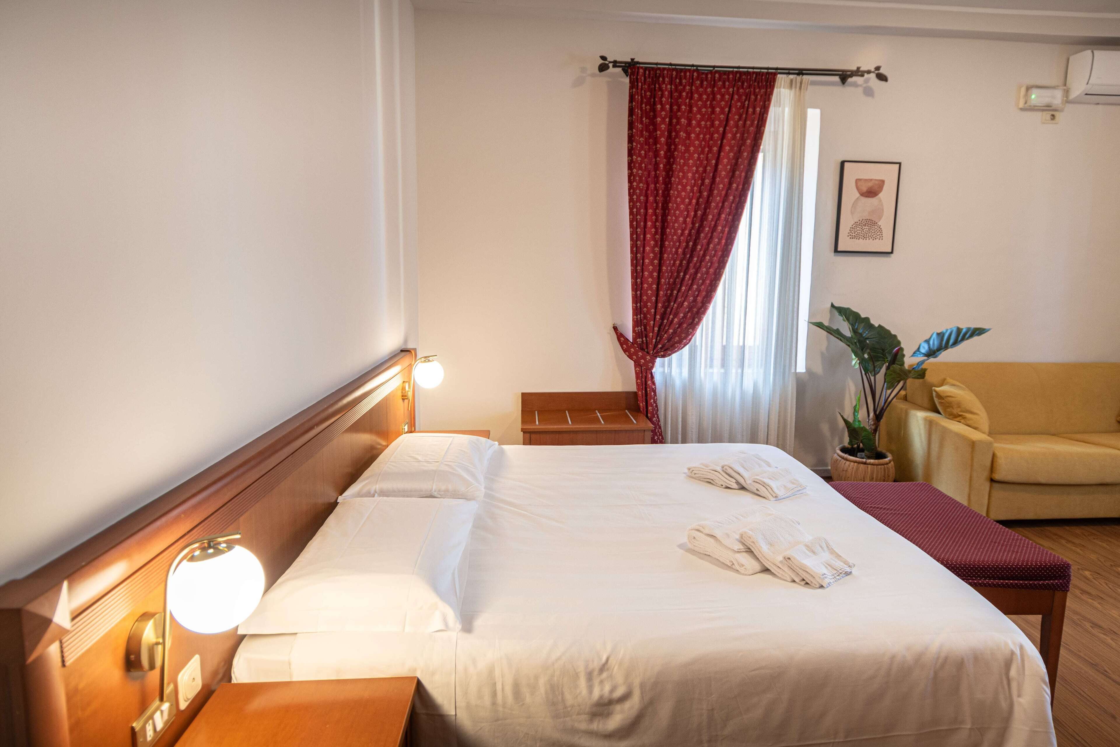 A hotel room with a bed, bedside table, lamp, and a potted plant.