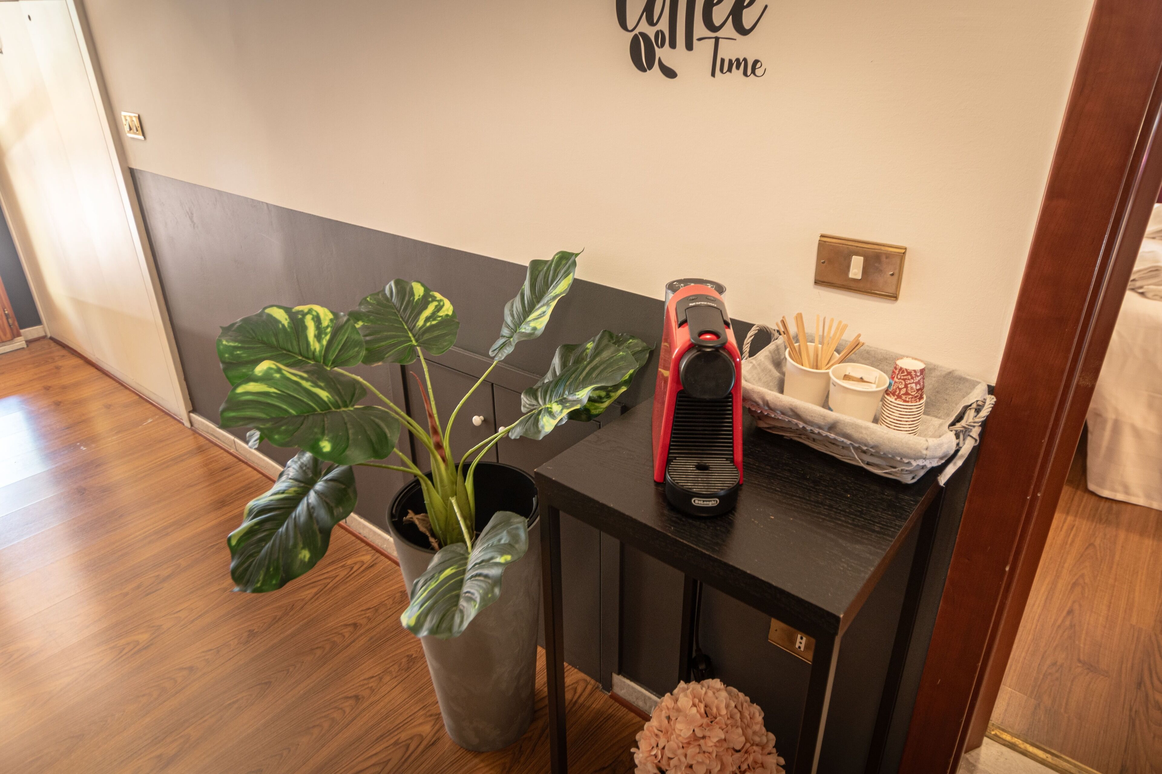 A room with a wooden floor, a potted plant, a small table with a coffee maker, and a wall with a 'Coffee Time' sign.