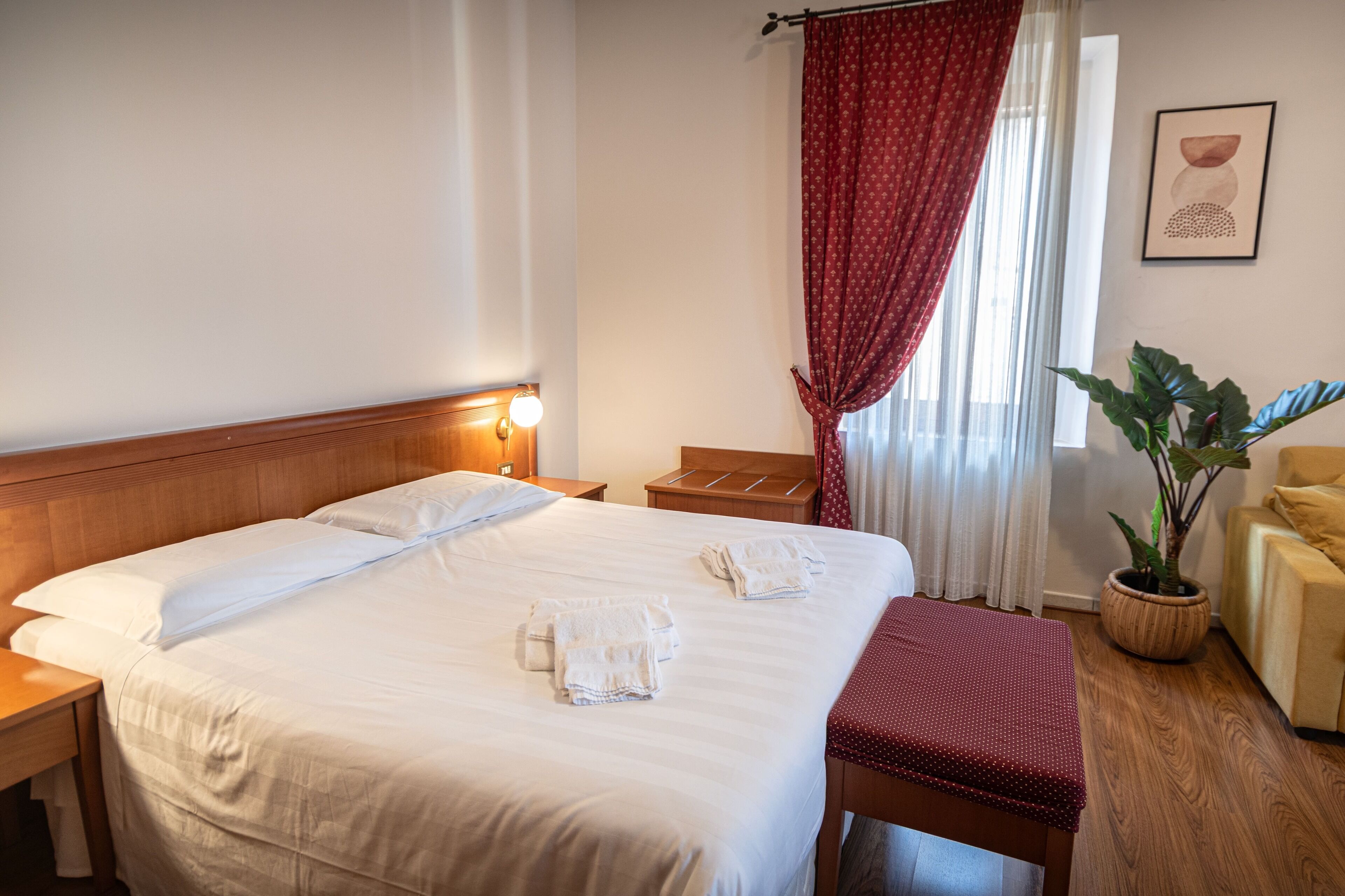 A hotel room with a large bed, a wooden headboard, a bedside table with a lamp, a small wooden nightstand, a red bench, a potted plant, and a framed picture on the wall.