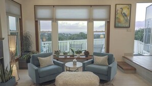 A living room with two blue armchairs, a white ottoman, a small round table, and a balcony with a railing.