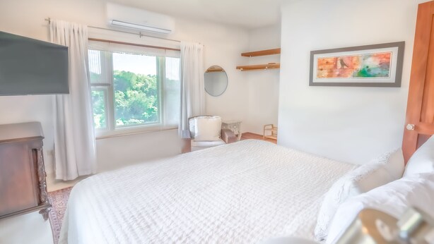 A bedroom with a large bed, a bedside table, a chair, a window with curtains, a wall-mounted mirror, and a framed picture.