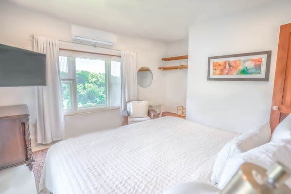 A bedroom with a large bed, a bedside table, a chair, a window with curtains, a wall-mounted mirror, and a framed picture.