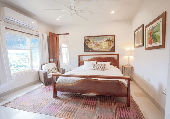 Une chambre avec un lit en bois, une grande fenêtre, un ventilateur de plafond et des œuvres d’art accrochées aux murs.