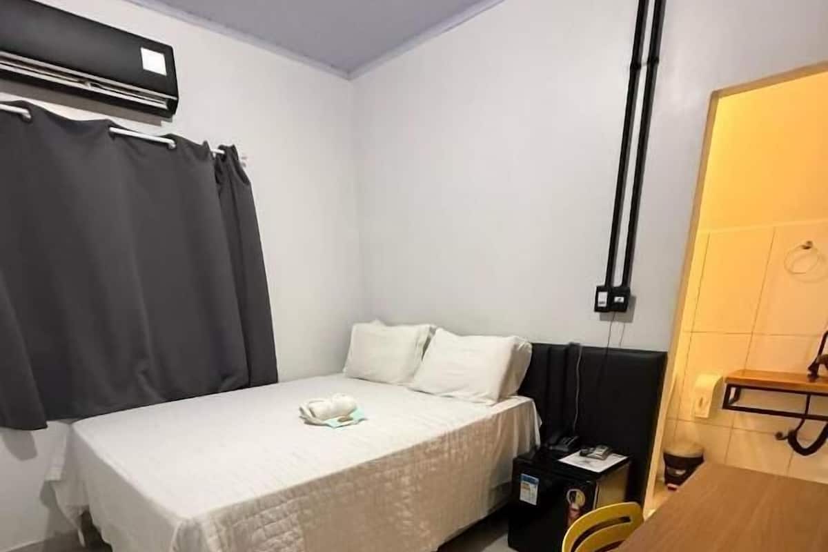 A small, clean room with a bed, a desk, and a chair. There is a window with curtains, a wall-mounted air conditioner, and a small table with a lamp.