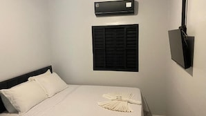 Una habitación de hotel con cama, aire acondicionado de pared, televisor y ventana con contraventanas.