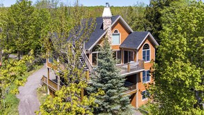 Une cabane en rondins à deux étages, dotée d’une cheminée en pierre, d’une terrasse en bois et d’une grande fenêtre.