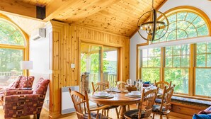 Un chalet en bois chaleureux, doté d’un coin repas avec une table ronde et des chaises, de grandes fenêtres offrant une vue sur la forêt et d’un plafond avec des poutres apparentes.