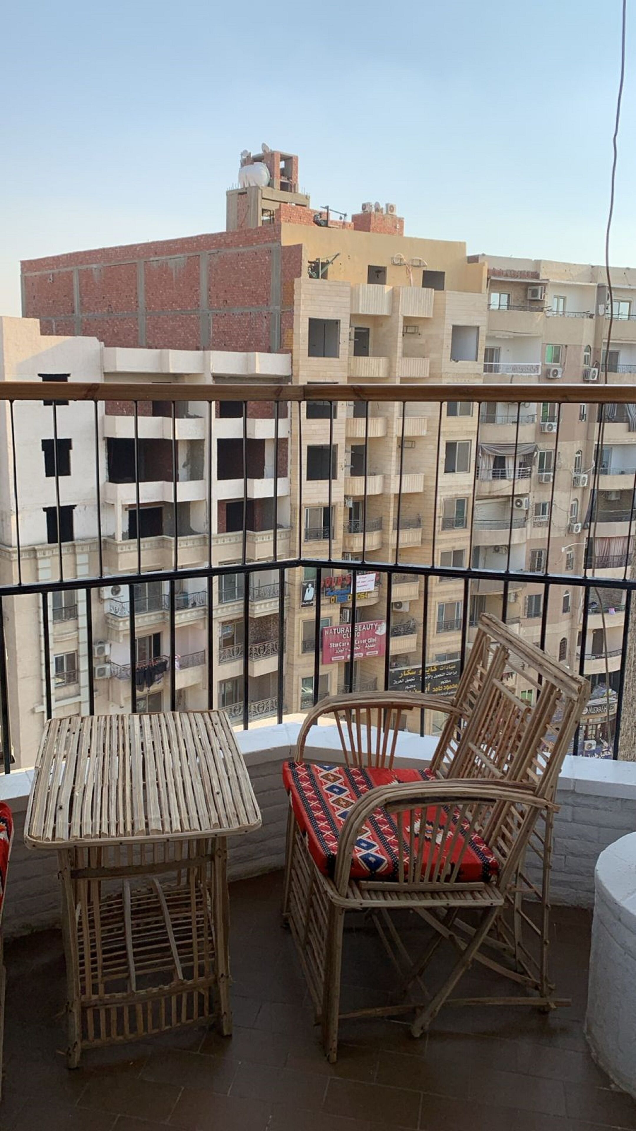 A balcony with a table and two wicker chairs, overlooking a residential area with multi-story buildings.
