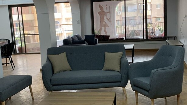 A spacious living area with a herringbone wooden floor, a central sofa, two armchairs, and a coffee table. There are large windows providing natural light and a view of the outside. - Pyramids Nights Hostel (Giza)