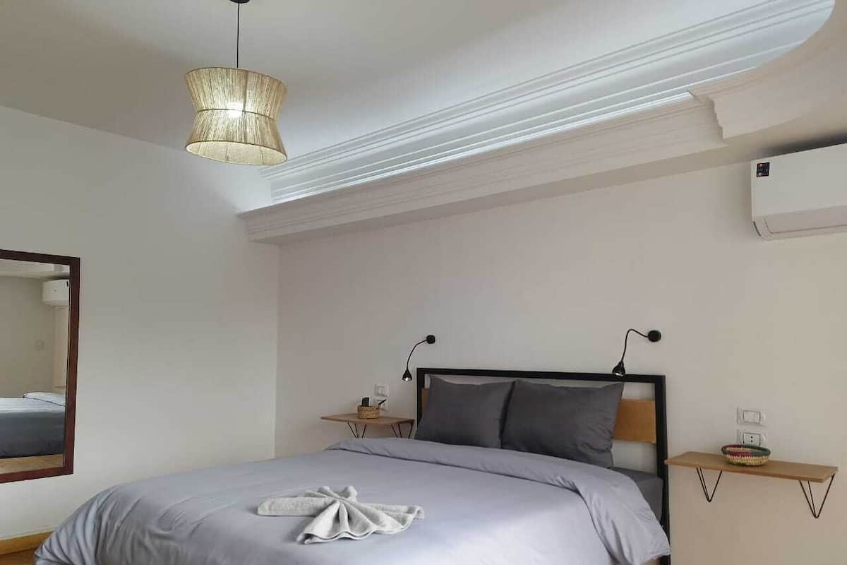 Un dormitorio con cama, mesitas de noche, lámpara colgante, espejo y equipo de aire acondicionado.