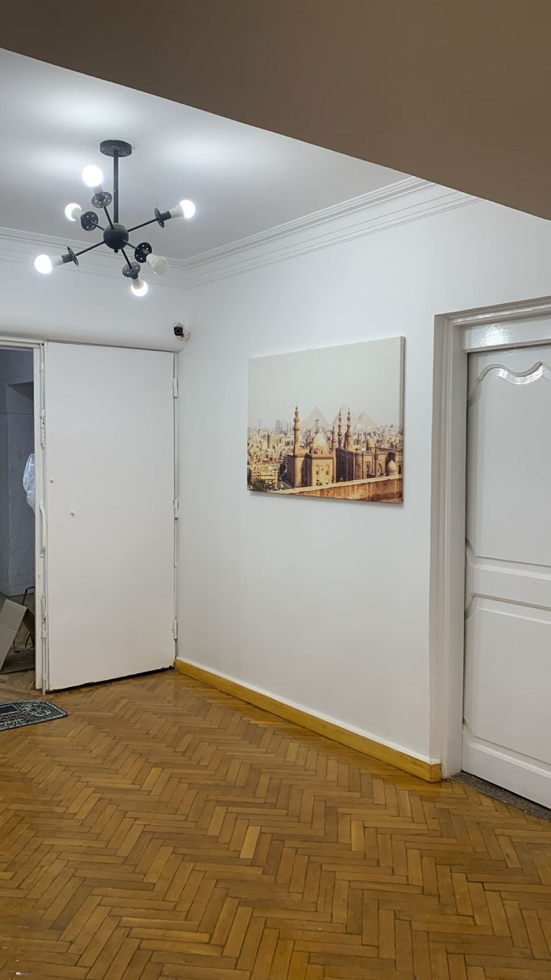 Una habitación con suelo de madera en dibujo de espiga, una puerta blanca, una pared blanca con un cuadro enmarcado y una lámpara en el techo.