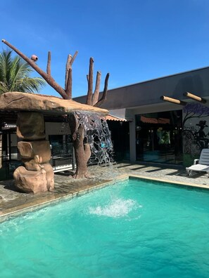 A pool with a waterfall feature and a stone structure resembling a tree trunk. - Pousada Aguas Quentes (Barra do Garcas)