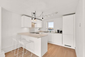 A modern kitchen with white cabinetry, a central island with bar stools, and a view through the window. - 2-Bed, 2-Bath Third Floor Flat (London)