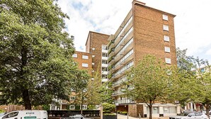 Un immeuble résidentiel à plusieurs étages, doté d’une façade en briques, de balcons et d’un espace de stationnement où sont garées des voitures.