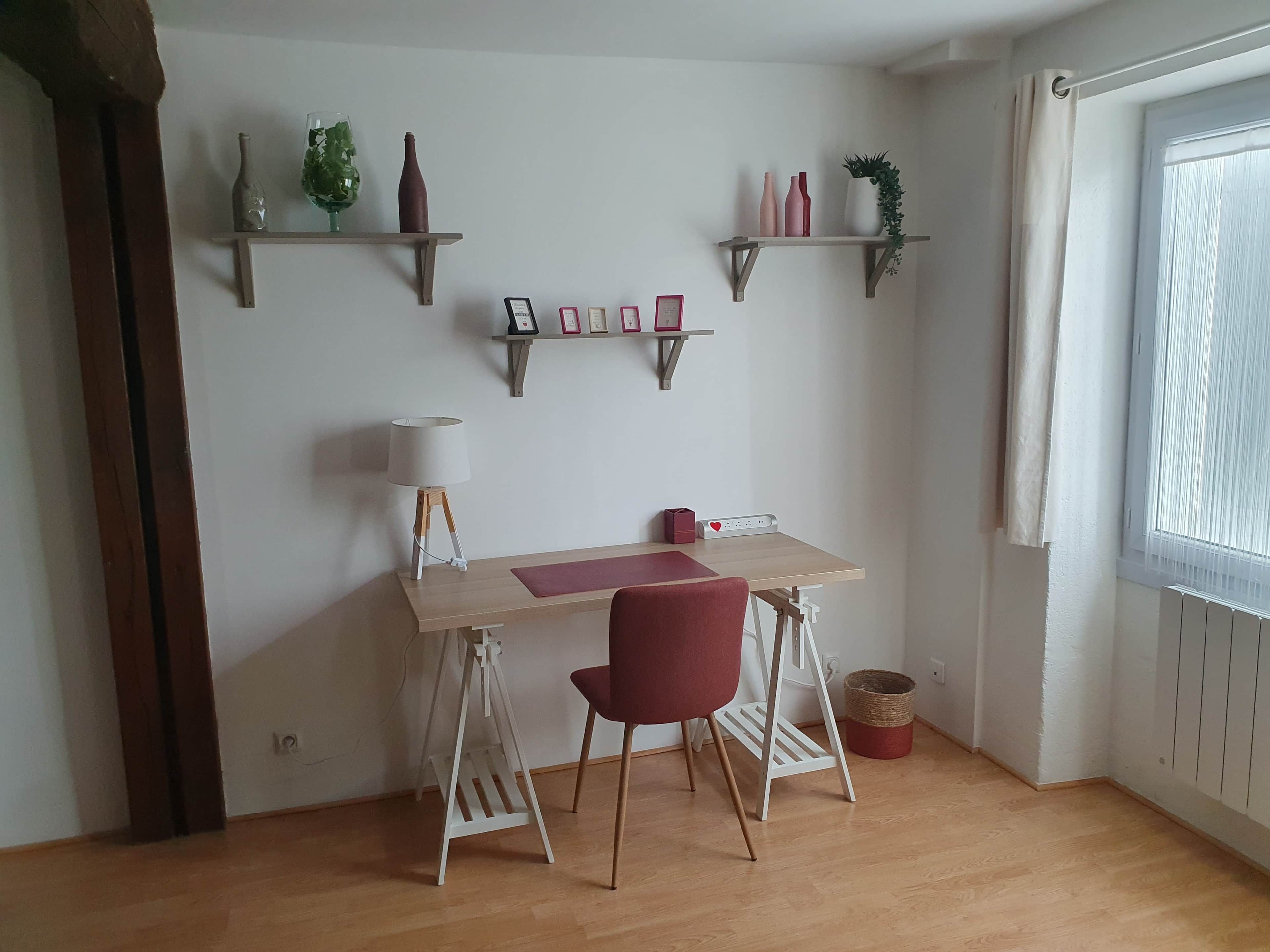 A room with a wooden desk, a red chair, a lamp, two shelves with decorative items, and a window with curtains.