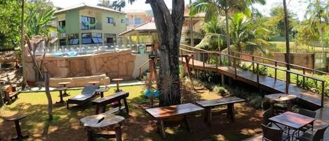 A wooden deck with tables and chairs, surrounded by lush greenery and a large tree.