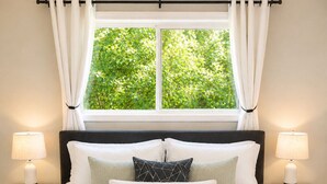 A bed with a white quilt, two bedside lamps, a window with curtains, and a view of greenery outside.