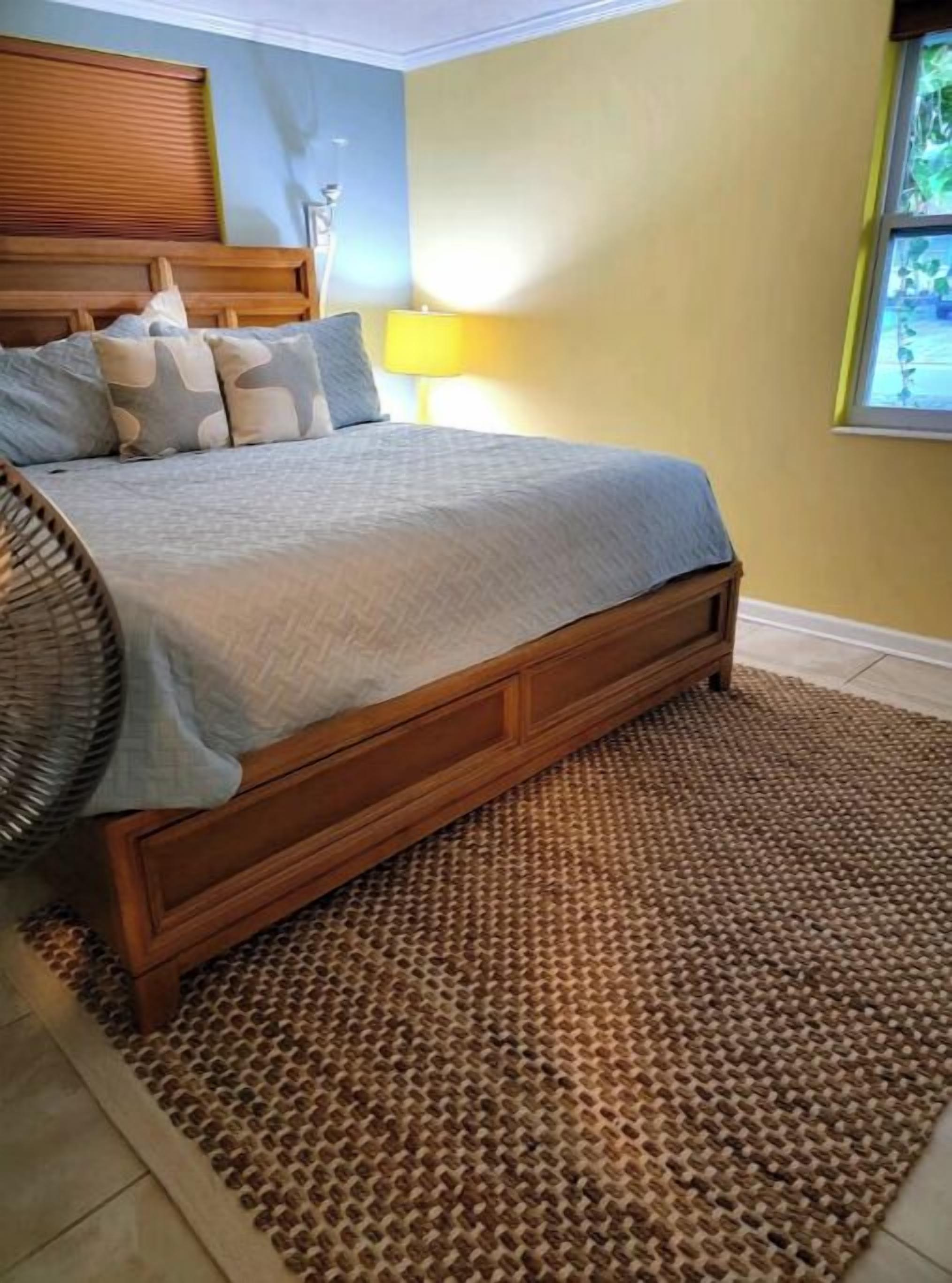 A bedroom with a wooden bed, a woven rug, and a window with a view of greenery.