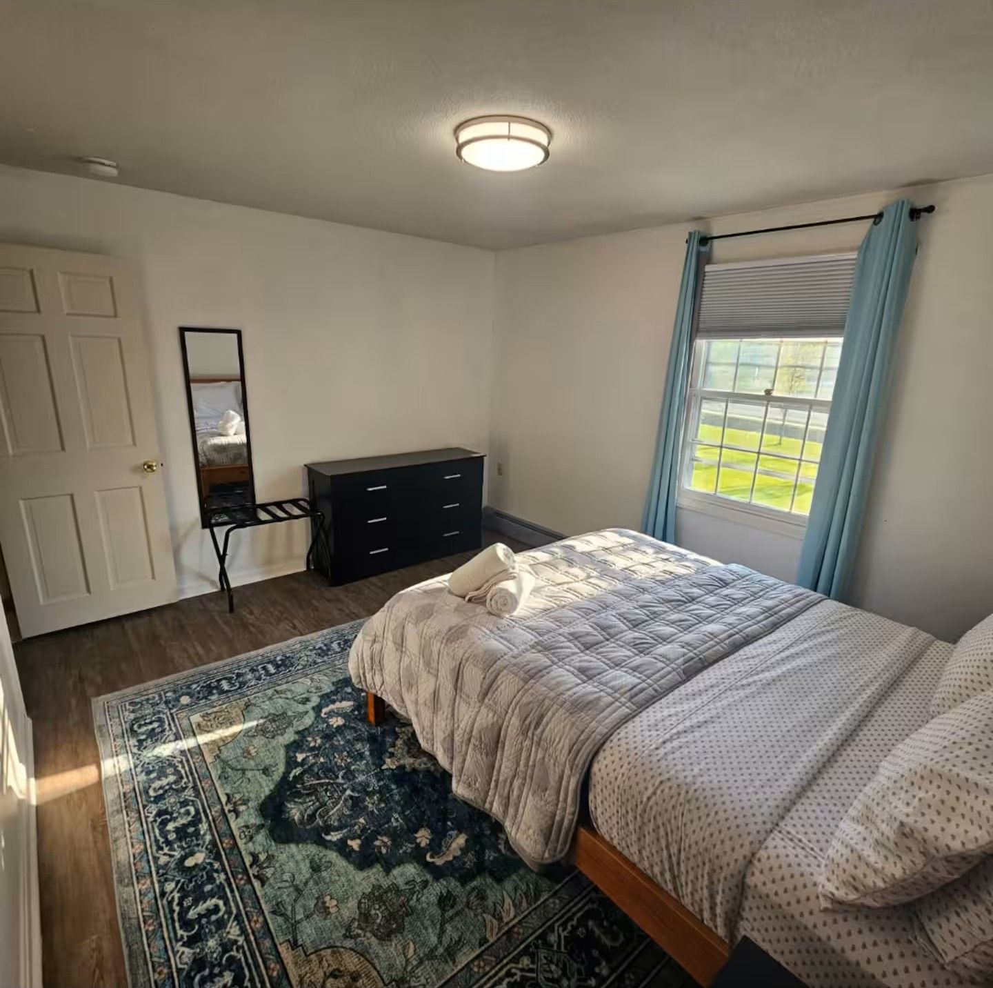 Une chambre à coucher avec un lit, une commode, un miroir, une fenêtre avec des rideaux et un tapis à motifs.