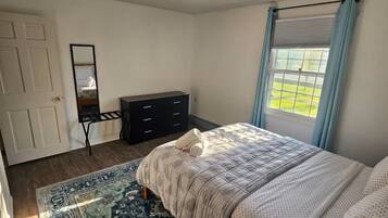 Une chambre à coucher avec un lit, une commode, un miroir, une fenêtre avec des rideaux et un tapis à motifs.