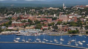 Un paisaje urbano costero con un puerto deportivo lleno de barcos, edificios históricos y una imponente aguja de iglesia al fondo.