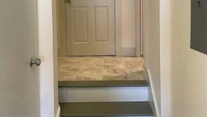 Un couloir étroit, avec une porte au fond, un interrupteur sur le mur et un sol carrelé.