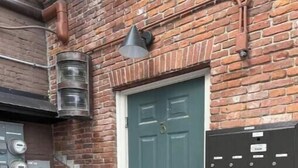 Un bâtiment en briques avec une porte verte, un compteur d’eau et un boîtier électrique noir.