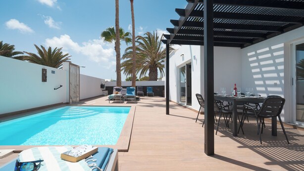 A pool area with lounge chairs and a table, surrounded by palm trees and a pergola.