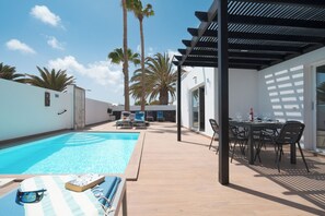 A pool area with lounge chairs and a table, surrounded by palm trees and a pergola. - Casa Afortunada, the pleasure of relaxation (Costa Teguise)